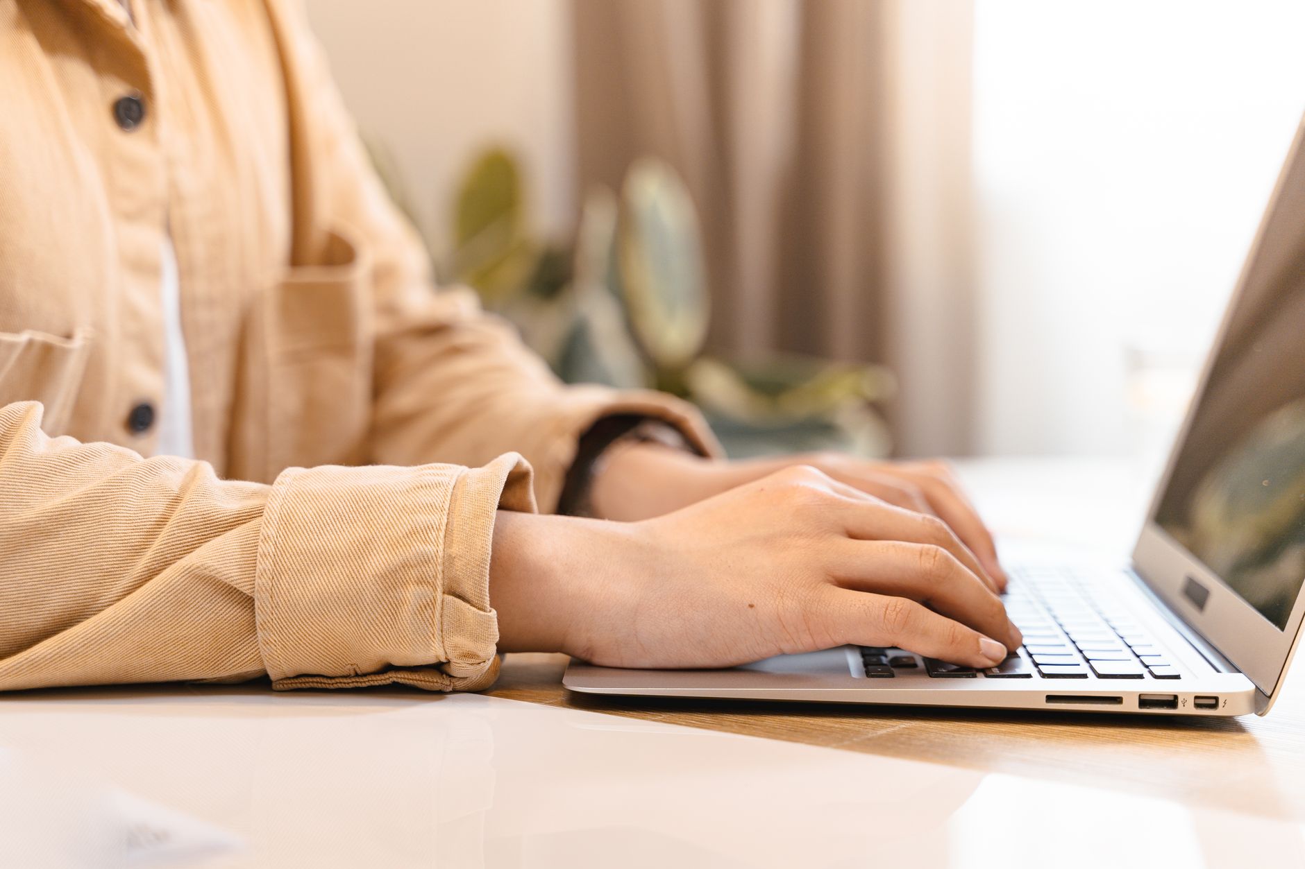 Close-up of hands typing on a laptop indoors, wearing a casual jacket.